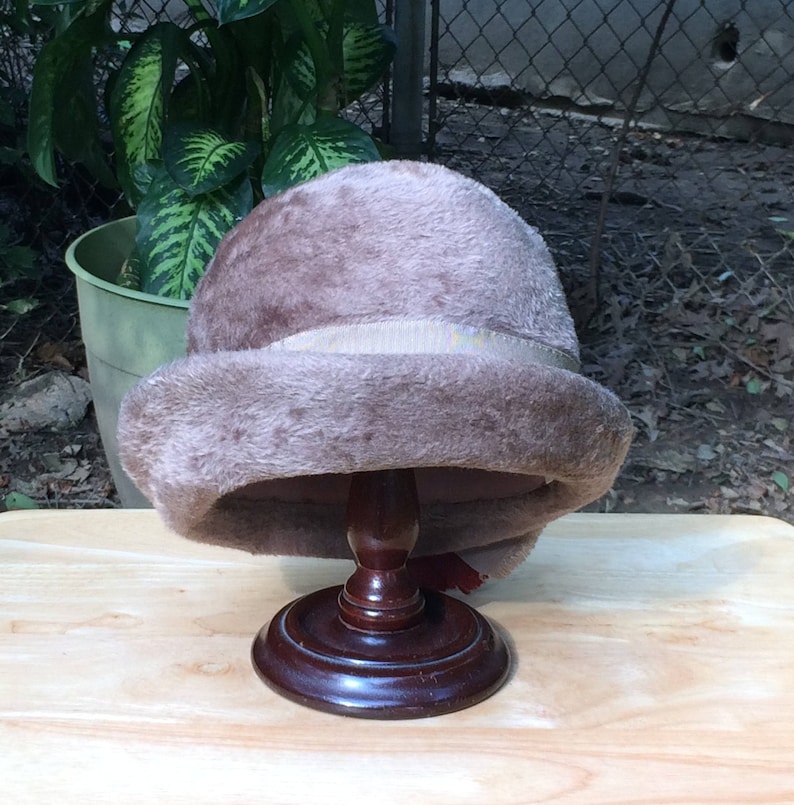 May include: A brown faux fur hat with a satin ribbon band. The hat is displayed on a wooden hat stand.