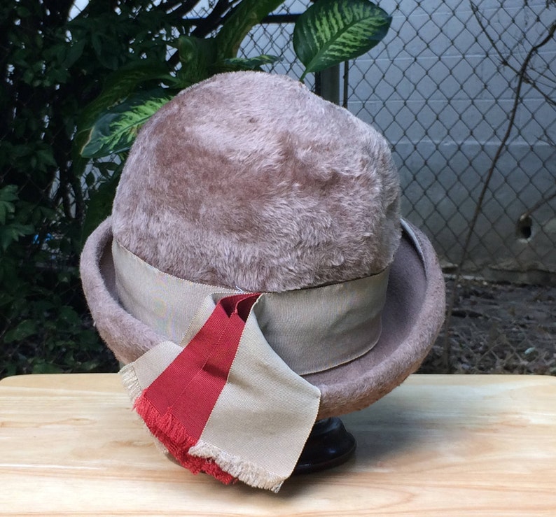May include: A brown felt hat with a wide brim and a beige ribbon. The hat has a red and beige ribbon tied around it.