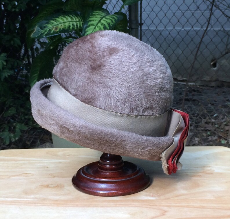 May include: A brown fur hat with a beige ribbon and a red and white ribbon trim. The hat is sitting on a wooden stand.