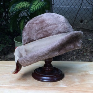 May include: A brown faux fur hat with a beige ribbon and a red trim. The hat is on a wooden display stand.