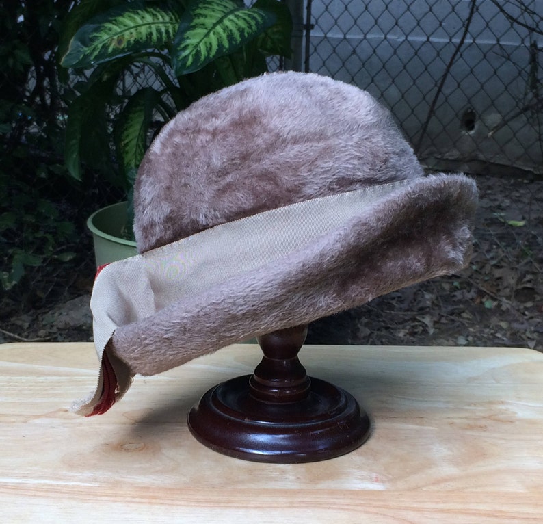 May include: A brown faux fur hat with a beige ribbon and a red trim. The hat is on a wooden display stand.