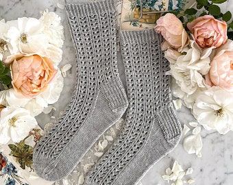 Peut inclure: Une paire de chaussettes grises en tricot avec un motif ajouré, suspendues à un cintre argenté. Les chaussettes sont sur une surface en marbre blanc avec des roses blanches et des pétales.