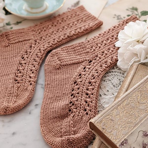 May include: A pair of knitted socks in a light brown color with a delicate lace pattern. The socks are lying on a marble surface with a book and flowers in the background.