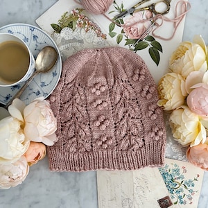 Könnte beinhalten: Ein altrosa gestrickter Beanie mit einem strukturierten Blatt- und Noppenmuster. Der Hut ist von Vintage-Artikeln umgeben, darunter eine Tasse Kaffee, antike Scheren und Postkarten, was eine gemütliche, handgefertigte Ästhetik schafft.
