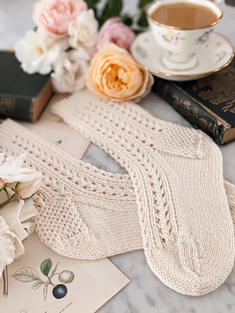 Peut inclure: Une paire de chaussettes en tricot cr&egrave;me avec des d&eacute;tails ajour&eacute;s. Les chaussettes sont pr&eacute;sent&eacute;es sur une surface en marbre avec des illustrations botaniques vintage, des roses et une tasse &agrave; th&eacute;. Les chaussettes ont un aspect textur&eacute; et un design classique.