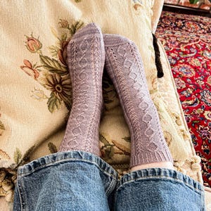 May include: A pair of light purple knitted socks featuring a vertical cable and lace pattern along the front. The socks have a ribbed cuff at the top and are worn with blue denim jeans. The knitted texture is visible throughout the fabric of the socks.