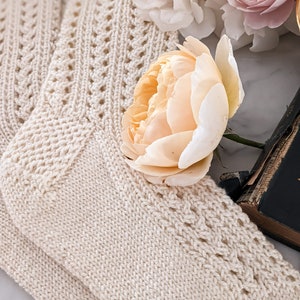 Peut inclure: Chaussettes en tricot cr&egrave;me avec un motif ajour&eacute; d&eacute;coratif. Les chaussettes sont pli&eacute;es et dispos&eacute;es avec une rose p&ecirc;che et d'autres fleurs blanches et roses. Un livre vintage est partiellement visible en arri&egrave;re-plan.