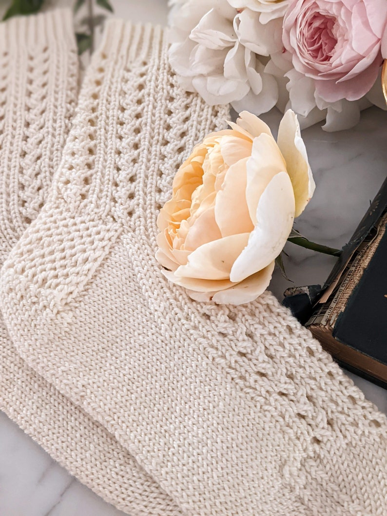 Peut inclure: Chaussettes en tricot cr&egrave;me avec un motif ajour&eacute; d&eacute;coratif. Les chaussettes sont pli&eacute;es et dispos&eacute;es avec une rose p&ecirc;che et d'autres fleurs blanches et roses. Un livre vintage est partiellement visible en arri&egrave;re-plan.