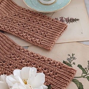 May include: Two pairs of brown knitted fingerless gloves with a delicate lace pattern, resting on a vintage book with botanical illustrations and white roses.
