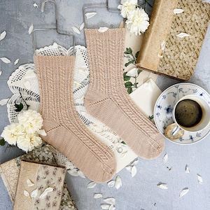 Könnte beinhalten: Zwei Paar beige gestrickte Socken, die an Drahtbügeln hängen. Die Socken haben ein Zopfmuster und werden auf einem grauen Hintergrund mit weißen Blütenblättern und alten Büchern präsentiert.
