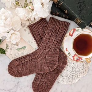 May include: A pair of brown knitted socks with a cable knit pattern. The socks are lying on a white marble surface with a cup of tea, a book, and flowers in the background.