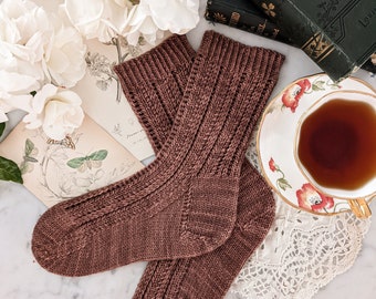 May include: A pair of brown knitted socks with a cable knit pattern. The socks are lying on a white marble surface with a cup of tea, a book, and flowers in the background.