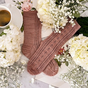 Könnte beinhalten: Ein Paar mauvefarbene Strickstrümpfe, einer uni und der andere mit einem Rautenmuster, werden zwischen weißen Blumen und Blumenkarten präsentiert. Eine Tasse Kaffee steht oben links auf einer weißen Untertasse. Die Socken sind ein gemütlicher, handgefertigter Artikel.