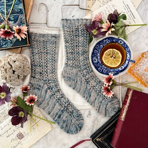 Puede incluir: Un par de calcetines tejidos a mano en un color azul y gris con un patrón de encaje. Los calcetines están colgados en una percha y están rodeados de libros, flores y una taza de té.