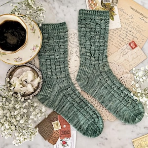 Könnte beinhalten: Ein Paar grüne Strickstrümpfe mit einem strukturierten Muster. Die Socken werden auf einer Marmoroberfläche mit Vintage-Postkarten, Spitze und einer Tasse Kaffee präsentiert.