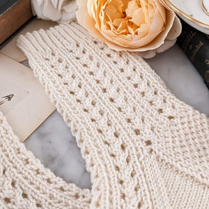 Peut inclure: &Eacute;charpe tricot&eacute;e de couleur cr&egrave;me avec un motif ajour&eacute; d&eacute;taill&eacute;. L'&eacute;charpe est faite d'un fil doux et textur&eacute;. L'arri&egrave;re-plan comprend une rose couleur p&ecirc;che, un livre vintage et une tasse &agrave; th&eacute;, sugg&eacute;rant une esth&eacute;tique vintage et confortable.