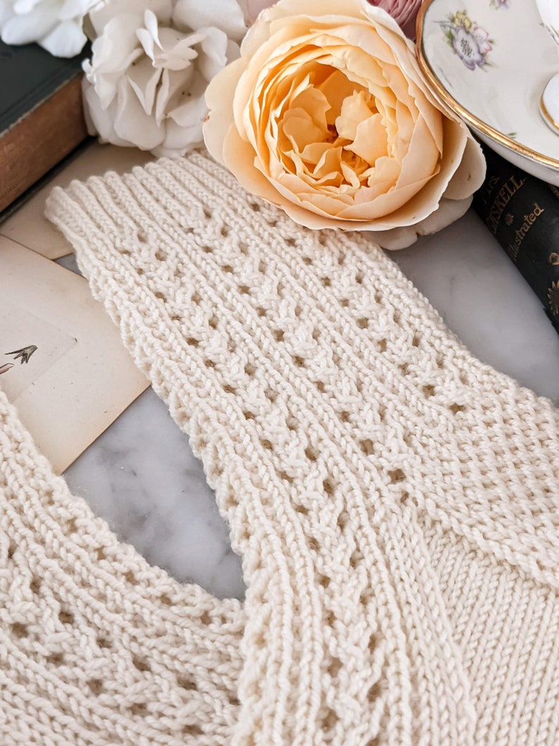 Peut inclure: &Eacute;charpe tricot&eacute;e de couleur cr&egrave;me avec un motif ajour&eacute; d&eacute;taill&eacute;. L'&eacute;charpe est faite d'un fil doux et textur&eacute;. L'arri&egrave;re-plan comprend une rose couleur p&ecirc;che, un livre vintage et une tasse &agrave; th&eacute;, sugg&eacute;rant une esth&eacute;tique vintage et confortable.