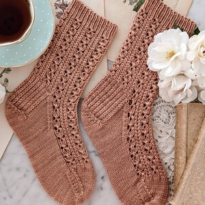 May include: A pair of brown knitted socks with a lacy pattern. The socks are lying on a white marble surface.