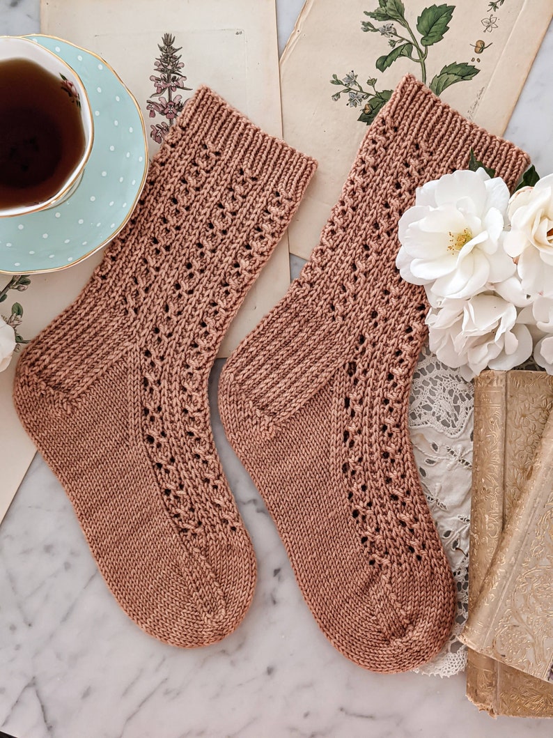 May include: A pair of brown knitted socks with a lacy pattern. The socks are lying on a white marble surface.