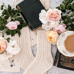 Peut inclure: Une paire de chaussettes en tricot cr&egrave;me avec un motif ajour&eacute; d&eacute;coratif, pr&eacute;sent&eacute;e avec des livres anciens, des estampes botaniques et des roses roses et cr&egrave;me. Une tasse et une soucoupe sont &eacute;galement pr&eacute;sentes.
