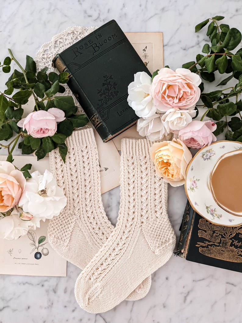 Peut inclure: Une paire de chaussettes en tricot cr&egrave;me avec un motif ajour&eacute; d&eacute;coratif, pr&eacute;sent&eacute;e avec des livres anciens, des estampes botaniques et des roses roses et cr&egrave;me. Une tasse et une soucoupe sont &eacute;galement pr&eacute;sentes.