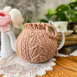 May include: A crocheted tea cozy in a light peach color with a delicate openwork pattern. The cozy is on a white teapot with a white lace doily underneath.