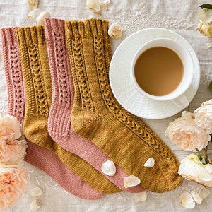 Può includere: Due paia di calzini lavorati a mano, giallo senape e rosa antico, con intricati motivi a treccia e pizzo. Una tazza bianca con caffè su un piattino, circondata da petali e fiori di rosa, su un tessuto bianco ricamato.
