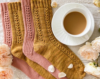Peut inclure: Deux paires de chaussettes tricotées à la main, jaune moutarde et rose poudré, avec des motifs de câbles et de dentelle. Une tasse blanche avec du café sur une soucoupe, entourée de pétales et de fleurs de roses, sur un tissu blanc brodé.