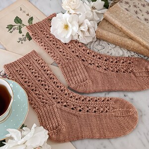 May include: A pair of knitted socks in a light brown color with a delicate lace pattern. The socks are lying on a vintage book with white roses and a teacup with a polka dot pattern.
