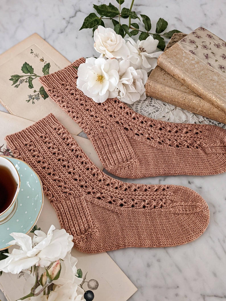 May include: A pair of knitted socks in a light brown color with a delicate lace pattern. The socks are lying on a vintage book with white roses and a teacup with a polka dot pattern.