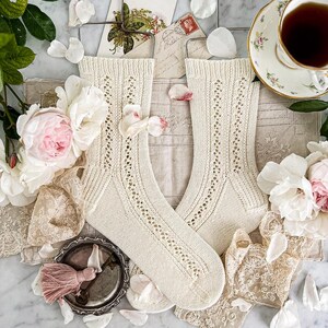 May include: A pair of cream-colored, knitted socks with a delicate openwork design, displayed on metal hangers. The socks are surrounded by pink and white flowers, lace, and a vintage teacup with tea.