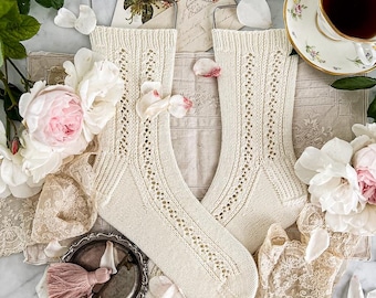 May include: Cream-coloured, knitted socks with a delicate openwork design, displayed on metal hangers. The socks are surrounded by pink and white flowers, lace, and a vintage teacup with tea.