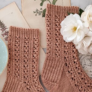 May include: A pair of knitted socks in a light brown color with a lacy pattern. The socks are lying on a white surface with a vintage book and white flowers.