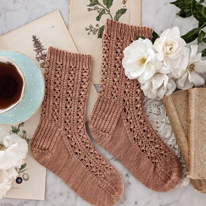 May include: A pair of brown knitted socks with a lacy pattern. The socks are lying on a marble surface with vintage books and flowers.