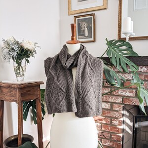 May include: A grey knitted scarf with a diamond pattern is displayed on a mannequin. The scarf is made of a thick yarn and has a textured appearance. The background includes a brick fireplace, framed artwork, and a vase of flowers.
