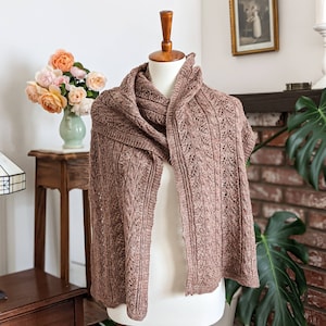 Puede incluir: Un chal de punto marrón con un patrón de punto de cable drapeado sobre un maniquí blanco. El chal se muestra contra una pared blanca con una chimenea de ladrillo y un jarrón de rosas rosas en el fondo.