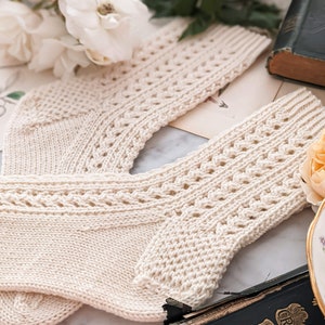 May include: Cream-coloured knitted socks with a decorative openwork pattern. The socks are displayed on a surface with flowers and a vintage book. The socks feature a ribbed cuff and a patterned design on the leg.