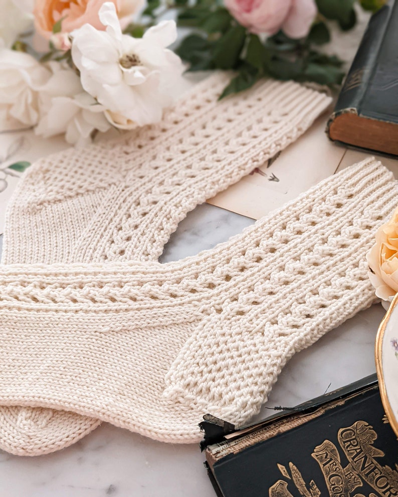 Peut inclure: Une paire de chaussettes en tricot cr&egrave;me avec un motif ajour&eacute; d&eacute;coratif. Les chaussettes sont pos&eacute;es sur une surface avec des fleurs et un livre ancien en arri&egrave;re-plan. Les chaussettes ont une bordure c&ocirc;tel&eacute;e et un motif sur la jambe.