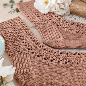May include: A pair of knitted socks in a light pink color with a delicate lace pattern. The socks are laid out on a white surface with white flowers.