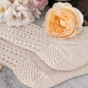 Peut inclure: Chaussettes en tricot cr&egrave;me avec un motif ajour&eacute; d&eacute;coratif. Les chaussettes sont pli&eacute;es et pos&eacute;es sur une surface en marbre, avec un bouquet de roses roses et p&ecirc;che en arri&egrave;re-plan. Une tasse et une soucoupe sont &eacute;galement visibles.