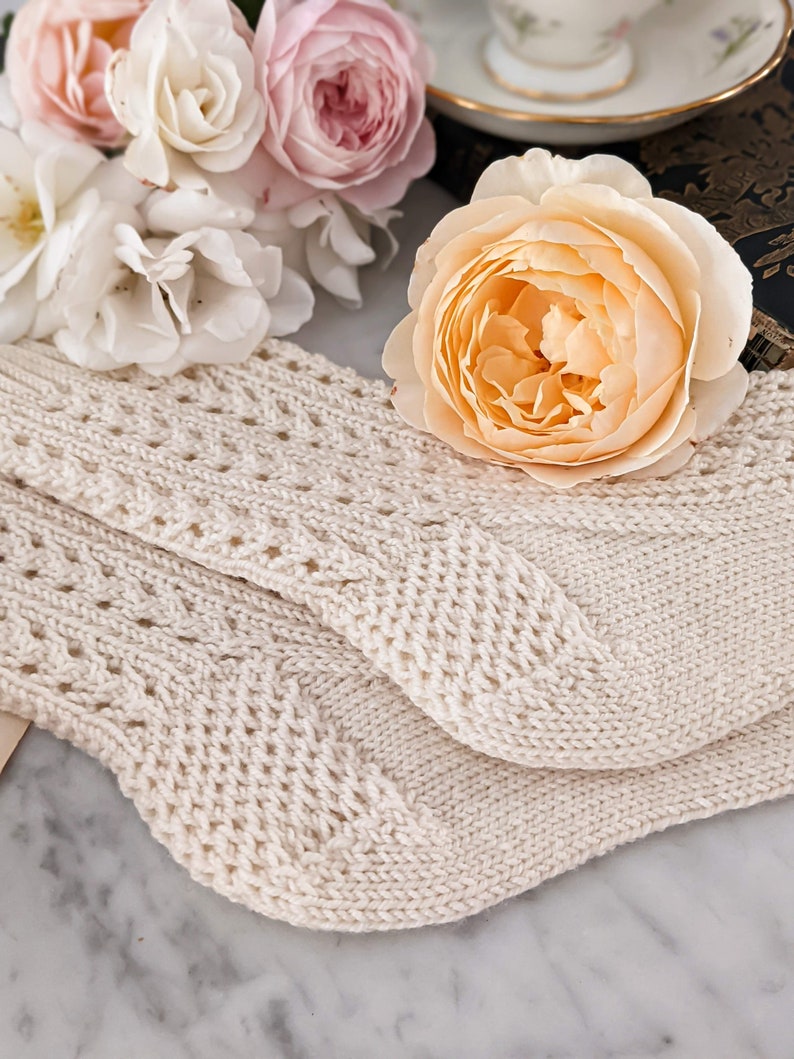 Peut inclure: Chaussettes en tricot cr&egrave;me avec un motif ajour&eacute; d&eacute;coratif. Les chaussettes sont pli&eacute;es et pos&eacute;es sur une surface en marbre, avec un bouquet de roses roses et p&ecirc;che en arri&egrave;re-plan. Une tasse et une soucoupe sont &eacute;galement visibles.