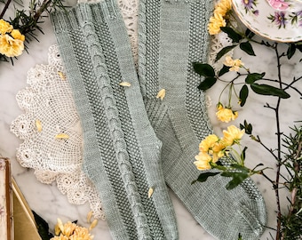 May include: A pair of sage green knitted socks with a cable knit pattern. The socks are lying on a white marble surface with a white doily and yellow flowers.