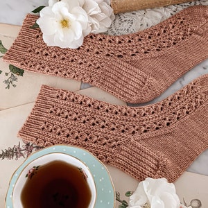 May include: A pair of hand-knitted socks in a dusty rose color with a delicate lace pattern. The socks are laid out on a table with a vintage book, a teacup, and white flowers.