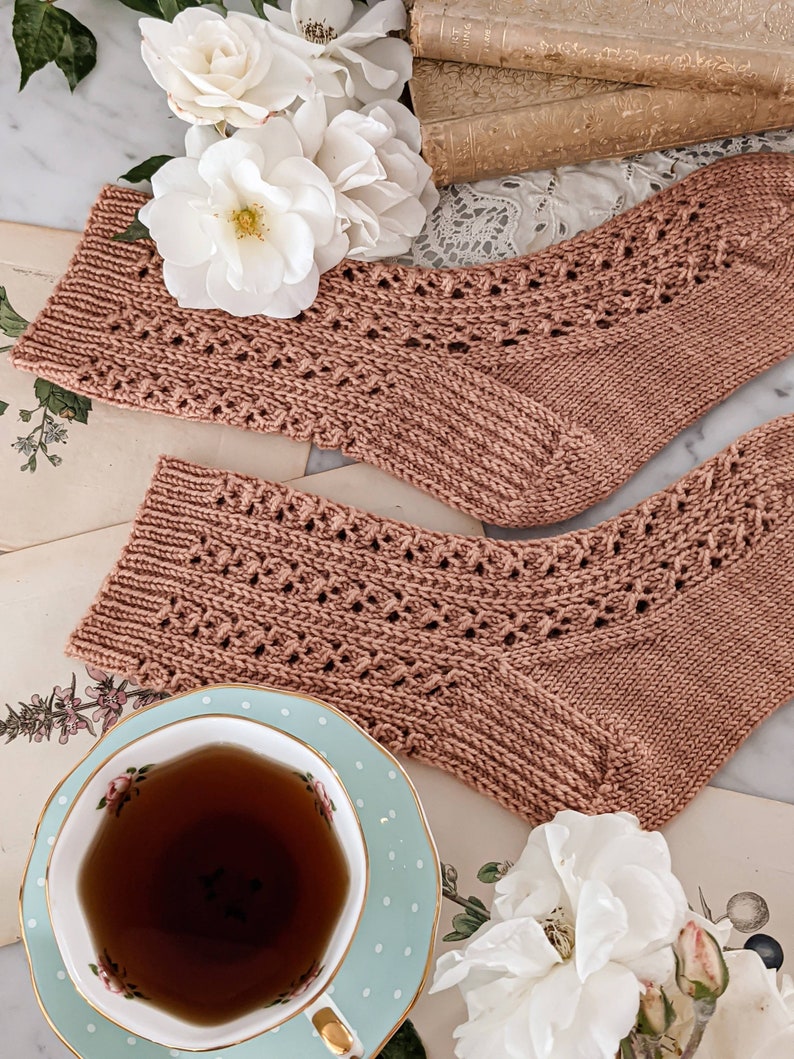 May include: A pair of hand-knitted socks in a dusty rose color with a delicate lace pattern. The socks are laid out on a table with a vintage book, a teacup, and white flowers.