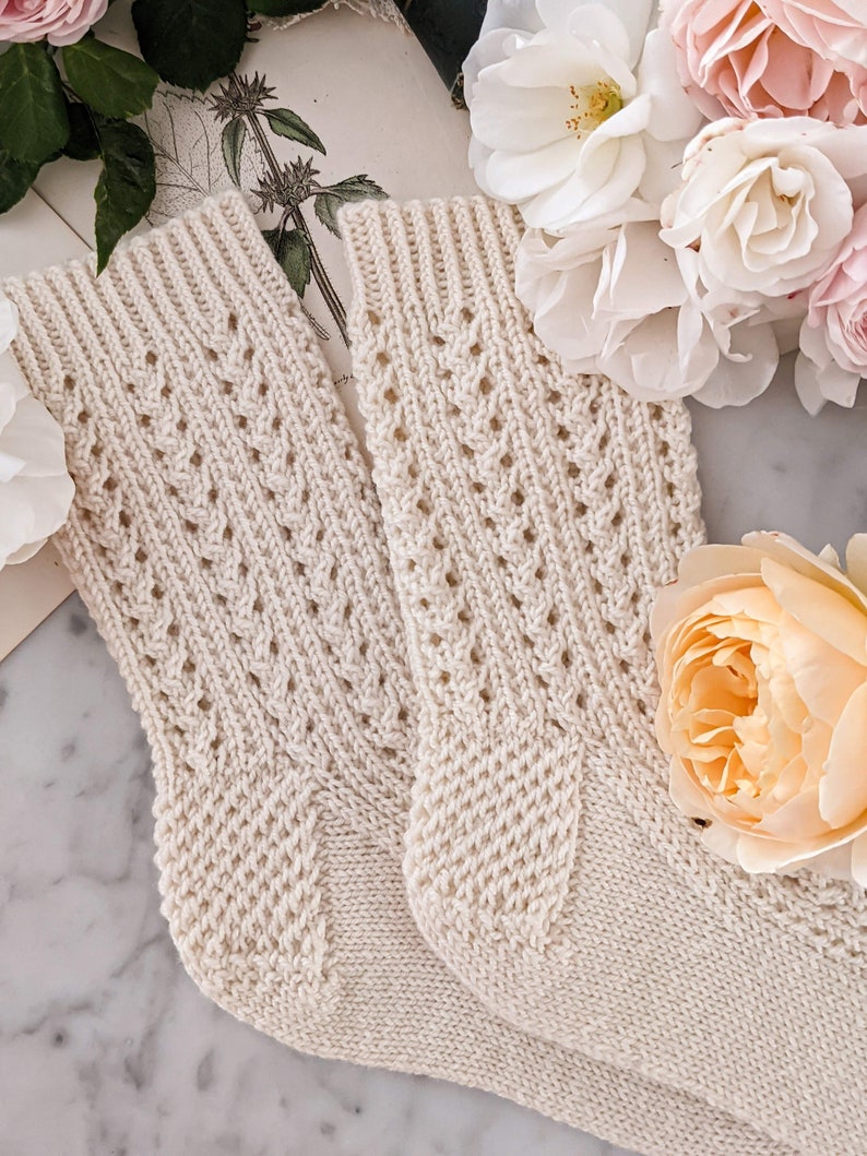 Peut inclure: Une paire de chaussettes en tricot cr&egrave;me avec un motif ajour&eacute; d&eacute;licat. Les chaussettes sont entour&eacute;es de roses roses et blanches, cr&eacute;ant une esth&eacute;tique chaleureuse et f&eacute;minine. Les chaussettes ont des poignets c&ocirc;tel&eacute;s et un design textur&eacute;.