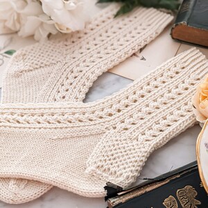 Peut inclure: Une paire de chaussettes en tricot cr&egrave;me avec un motif ajour&eacute; d&eacute;coratif. Les chaussettes sont pr&eacute;sent&eacute;es sur une surface en marbre avec un livre ancien et des fleurs en arri&egrave;re-plan. Les chaussettes ont un bord c&ocirc;tel&eacute; et un design d&eacute;taill&eacute;.