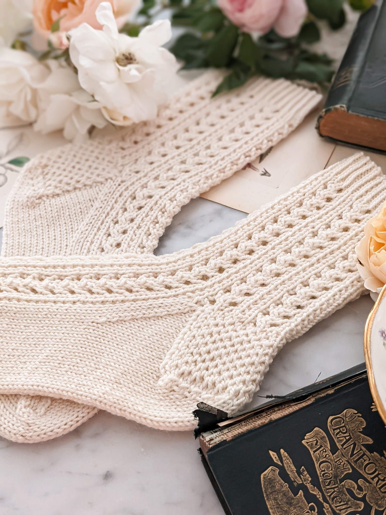 Peut inclure: Une paire de chaussettes en tricot cr&egrave;me avec un motif ajour&eacute; d&eacute;coratif. Les chaussettes sont pr&eacute;sent&eacute;es sur une surface en marbre avec un livre ancien et des fleurs en arri&egrave;re-plan. Les chaussettes ont un bord c&ocirc;tel&eacute; et un design d&eacute;taill&eacute;.