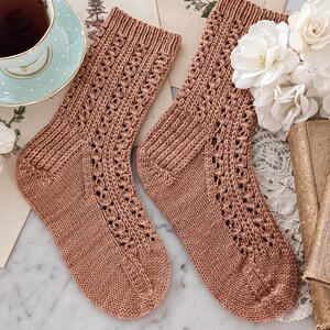 May include: A pair of brown knitted socks with a delicate lace pattern. The socks are laid out on a marble surface with vintage books and flowers.