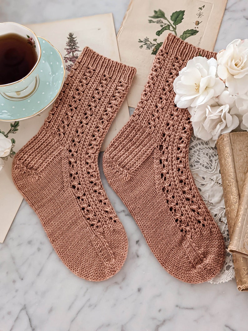 May include: A pair of brown knitted socks with a delicate lace pattern. The socks are laid out on a marble surface with vintage books and flowers.