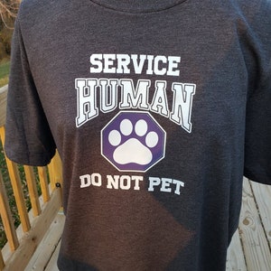 May include: A dark gray t-shirt with the text "SERVICE HUMAN" in white letters and a purple paw print inside a hexagon. The text "DO NOT PET" is below the paw print.